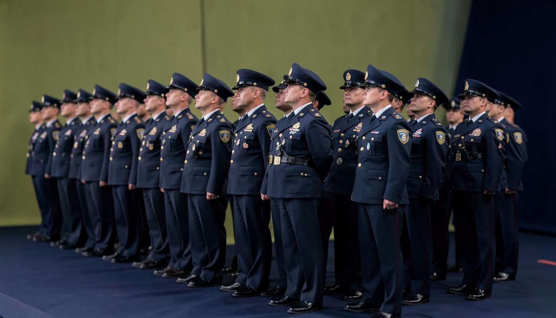 A row of uniformed personnel standing at attention, with a soft olive and navy backdrop, conveying disciplined service recruitment.