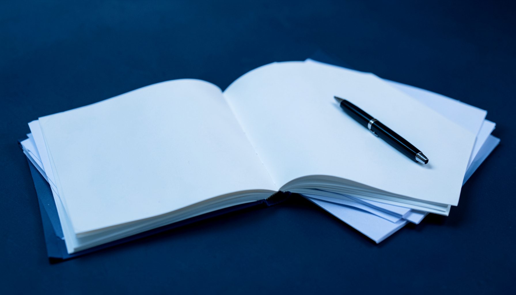 Soft blue and dark navy toned image of an open notebook and pen beside stacked papers, suggesting quiet study and exam preparation